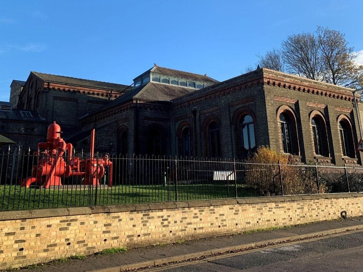 A large red water pump on display in the gardens of the Cambridge Museum of Technology.