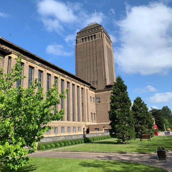 Front view of Cambridge University Library, a striking 20th-century brick building with a tall central tower, surrounded by greenery.