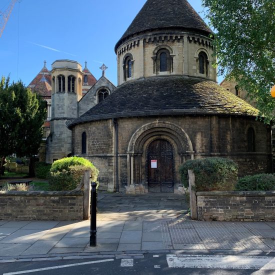 Exterior view of the Round Church in Cambridge, a distinctive Norman-style building with a circular nave and stone facade, set against a bright sky.